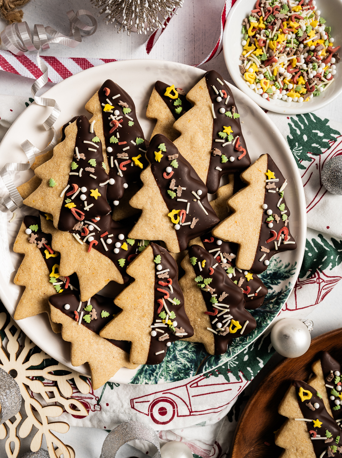 tree shaped cinnamon shortbread cookies half dipped in chocolate with sprinkles bowl of sprinkles ornaments red and white striped ribbon