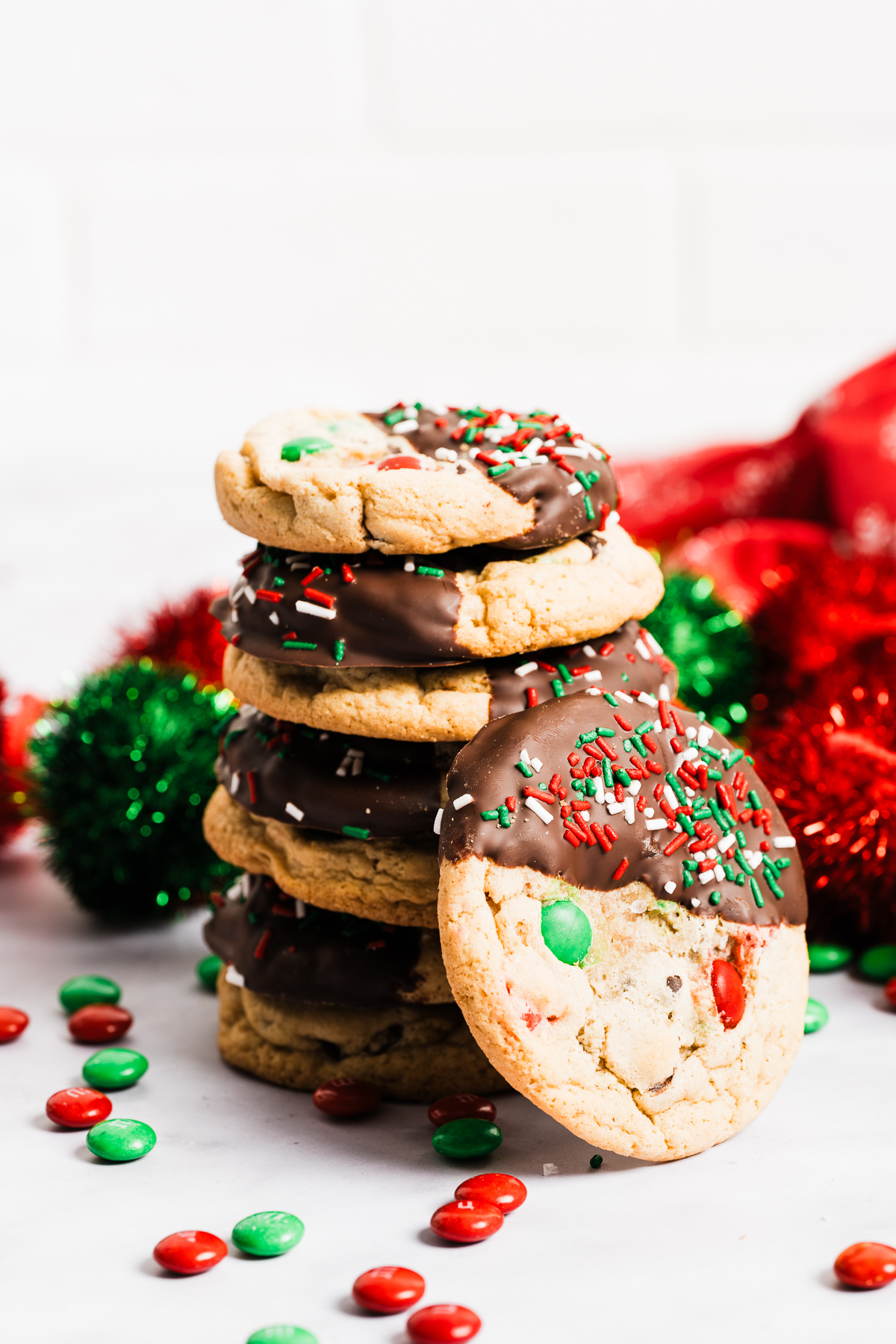 red and green M&M chewy chocolate chip cookies half dipped in chocolate decorated with red white and green sprinkles in a stack with one on its side