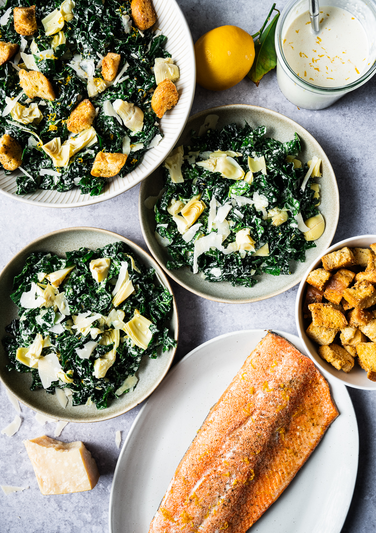 Kale Caesar salads in bowls platter of roasted salmon bowl of croutons jar of Caesar dressing